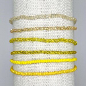 May include: A set of six beaded bracelets arranged in a vertical stack. The bracelets feature a gradient of yellow tones, ranging from a translucent pale yellow at the top to a solid, opaque bright yellow at the bottom, all made of small beads.