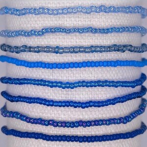 May include: A collection of eight beaded bracelets in varying shades of blue. The bracelets are made of small, round beads and are displayed on a white fabric background. The colors range from light sky blue to deep navy.