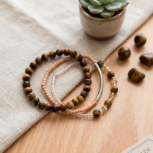 May include: Three beaded bracelets featuring tiger eye stones, clear beads, and peach-coloured beads. A small succulent plant in a ceramic pot and several tiger eye stones are also visible. The bracelets are on a wooden surface.