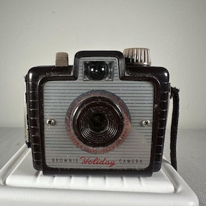 May include: A vintage Brownie Holiday camera with a dark brown plastic body and a silver front faceplate featuring horizontal lines. The lens is surrounded by red text reading Made in Rochester, N.Y. U.S.A. by Eastman Kodak Company.