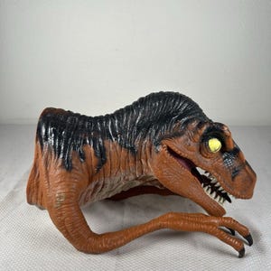 May include: A brown and black dinosaur hand puppet featuring a textured skin pattern, a wide open mouth with visible white teeth, and bright yellow eyes. The puppet is designed to fit over a hand, showing the head and two long, clawed arms.