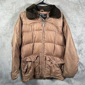 Puede incluir: Chaqueta acolchada marrón con cuello de piel sintética marrón oscuro. La chaqueta tiene cierre de cremallera, dos bolsillos delanteros y la marca "Bailey" es visible en una etiqueta. La chaqueta está colgada de una percha negra.