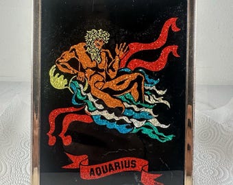 May include: A rectangular framed wall art piece featuring a glittery illustration of a figure pouring water, set against a black background. The design includes red ribbons and the word AQUARIUS printed in black on a red banner at the bottom.