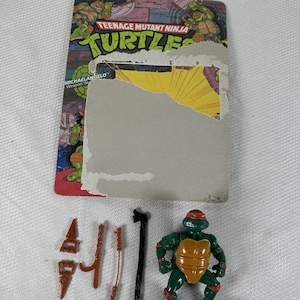 May include: A Teenage Mutant Ninja Turtle action figure of Michelangelo, with orange mask and shell, and various weapons. The figure is green, with an orange belt and mask. The packaging is visible behind the figure.
