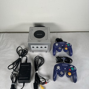 Può includere: Una console Nintendo GameCube color argento con coperchio del disco circolare nero, due controller cablati viola con pulsanti multicolori, un alimentatore nero e un set di cavi AV gialli, bianchi e rossi per la connessione video e audio.