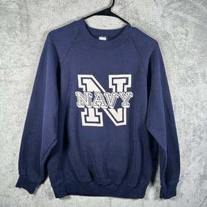 Könnte beinhalten: Marineblaues Sweatshirt mit dem Wort "NAVY" in weißen Blockbuchstaben. Das Sweatshirt hat lange Ärmel und einen Rundhalsausschnitt. Das Kleidungsstück hängt an einem schwarzen Kleiderbügel vor grauem Hintergrund.