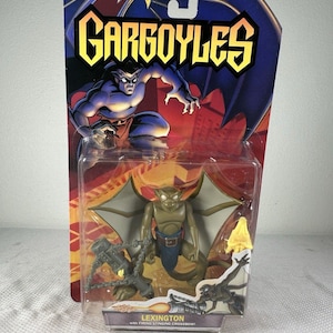 Puede incluir: Una figura de acción de Lexington de Gargoyles en su empaque blíster original. La figura es de color canela con alas grandes y un taparrabos azul, acompañada de un accesorio de ballesta gris. La tarjeta muestra el logo de Gargoyles.