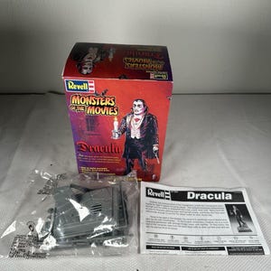 Peut inclure: Un kit de modèle réduit Revell Monsters of the Movies Dracula dans une boîte rouge et violette illustrée de Dracula tenant une bougie. L'ensemble comprend un sachet plastique transparent contenant des pièces en plastique gris et une notice.