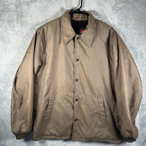 Puede incluir: Chaqueta de entrenador color marrón claro con cierre de botones, dos bolsillos delanteros y cuello y puños marrón oscuro. La chaqueta está colgada de una percha negra sobre un fondo gris.