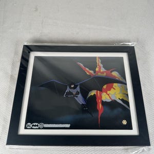 Könnte beinhalten: Ein gerahmtes Animations-Cel, das Batman in einem grauen und schwarzen Anzug mit Umhang zeigt, wie er vor einem dunklen Hintergrund mit einer großen roten und gelben Explosion gleitet. Der Rahmen ist schwarz mit weißem Passepartout und DC-Logo.