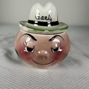 May include: A ceramic garlic-shaped container with a smiling face and a hat. The hat is white with a green brim and the word "Garlic" written on it. The container is pink with red cheeks.