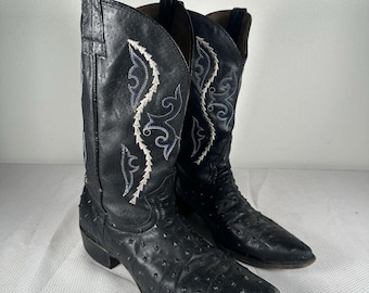 May include: A pair of black cowboy boots with a textured surface. The boots feature decorative white and blue stitching along the shaft. The boots have pointed toes and a low heel.