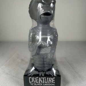 May include: A grey Creature from the Black Lagoon bubble wand bottle encased in clear plastic packaging. The base features the text "CREATURE FROM THE BLACK LAGOON BUBBLE-WAND BOTTLE" in white lettering against a black background with a graphic burst.