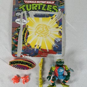 May include: A Teenage Mutant Ninja Turtles action figure, still on its original packaging. The figure is a green turtle with a red and blue torso, and comes with accessories. The packaging features the words "Teenage Mutant Ninja Turtles" and "Kowabunga!"