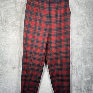 May include: Red and black plaid pants hanging on a black hanger. The pants have a classic tartan pattern with a button closure and pleats at the waist. The fabric appears to be a medium-weight material.