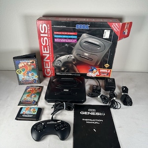 Może przedstawiać: Zestaw konsoli do gier wideo Sega Genesis zawierający czarną konsolę, przewodowy kontroler, zasilacz i kable AV. Zestaw zawiera kartridż z grą Sonic the Hedgehog 2, oryginalne etui, instrukcję obsługi oraz oryginalne pudełko.