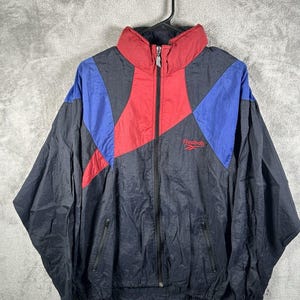 May include: A vintage Reebok windbreaker jacket in black, red, and blue. The jacket features a full zip, two front pockets, and the Reebok logo on the chest. The collar is red, with blue and red geometric shapes on the shoulders.