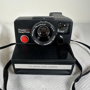 May include: Black Polaroid Pronto! BC RangeFinder camera with a red button and a black strap. The camera has the text "POLATRIPLET" and "POLAROID LAND CAMERA" printed on it. The camera is a vintage instant film camera.