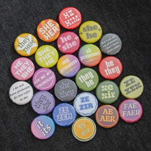 May include: A collection of colourful round buttons with various pronoun options, including "they/them", "she/her", "he/him", "xe/xem", "ze/zir", "ae/aer", "fae/faer", "ey/eir", and "it/its".