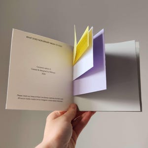 May include: An open book with the title "WHAT DOES NON-BINARY MEAN TO YOU?" visible. The book has yellow and purple pages that are partially open. The text "Container edition 5 Created & designed by fithmars 2025" is also visible.