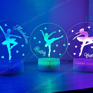 May include: Three white round night lights with a ballerina silhouette and a personalized name. The names are Madeline, Betty, and Veronica. The lights are illuminated in different colors, blue, green, and purple.