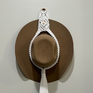 May include: White macrame hat holder with a brown felt hat hanging from it. The holder has a wooden bead at the top and a tassel at the bottom.