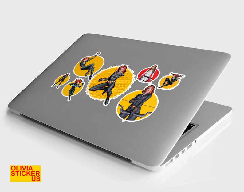 May include: A silver laptop computer with six yellow and red circular stickers featuring a female superhero character in a black suit. The stickers are arranged in a pattern on the laptop's lid.