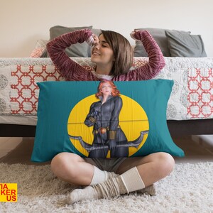 May include: A teal blue pillow with a yellow circle and a black and red illustration of a woman holding a bow and arrow. The woman is wearing a black outfit with a red belt and a red headband. The pillow is on a white rug.