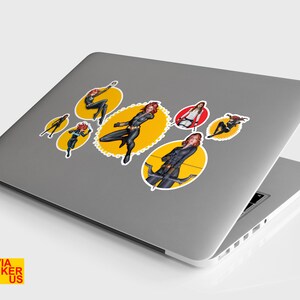 May include: A silver laptop computer with six yellow and red circular stickers featuring a female superhero character in a black suit. The stickers are arranged in a pattern on the laptop's lid.
