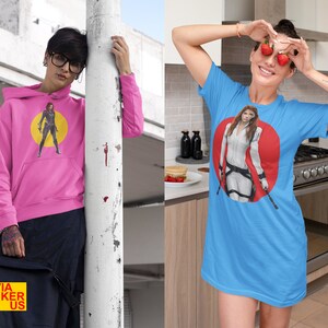 May include: Two women wearing t-shirts with a graphic of a female superhero. The woman on the left is wearing a pink hoodie with a yellow circle around the graphic. The woman on the right is wearing a light blue t-shirt with a red circle around the graphic. Both shirts feature the text "Olivia Sticker US".
