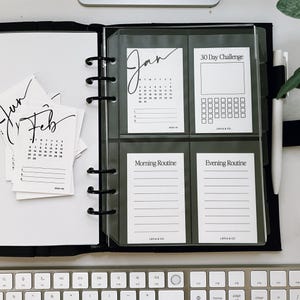 May include: A black binder with clear plastic pockets containing printable planner pages. The pages include a January calendar, a 30-day challenge tracker, a morning routine log, and an evening routine log. The pages are labeled with the text "Jan", "30 Day Challenge", "Morning Routine", and "Evening Routine".