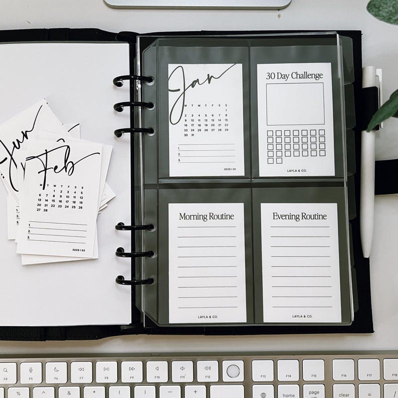 May include: A black binder with clear plastic pockets containing printable planner pages. The pages include a January calendar, a 30-day challenge tracker, a morning routine log, and an evening routine log. The pages are labeled with the text "Jan", "30 Day Challenge", "Morning Routine", and "Evening Routine".