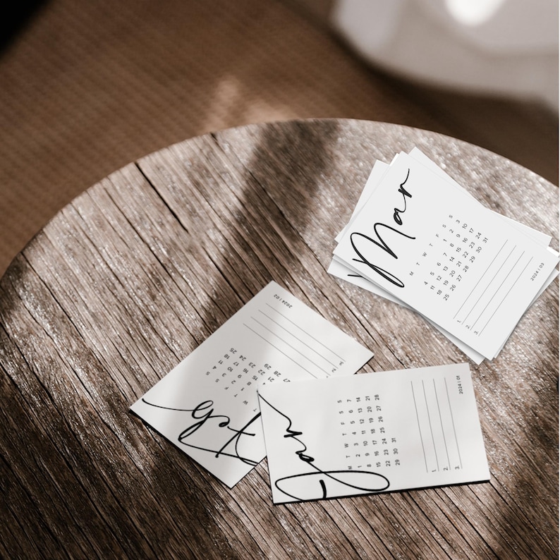 May include: Three white printable calendar cards with black handwritten text. The cards are for the months of January, February, and March. The cards are on a wooden surface.