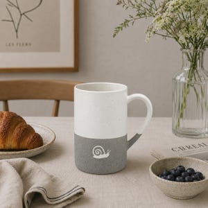 May include: A two-toned ceramic mug featuring a speckled white upper section and a solid grey lower section. A small white snail illustration is centered on the grey portion. The mug has a curved handle and a cylindrical shape with a smooth finish.