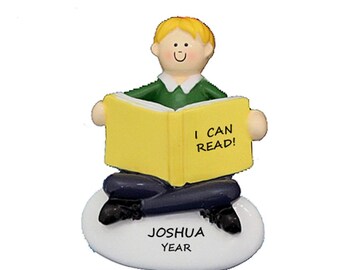 May include: A figurine of a child with blond hair wearing a green shirt and blue pants, reading a yellow book that says "I CAN READ!". The figurine is sitting on a white oval base with the text "JOSHUA YEAR".