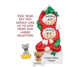 May include: A personalized Christmas ornament featuring two figures in red suits with green accents. The names "Megan" and "Travis" are displayed. A dog and cat figurine are included, with the name "Aspen" on a bone. Text reads "Pick your pet you would like us to add from our large selection" and "Christmas 'Year'".