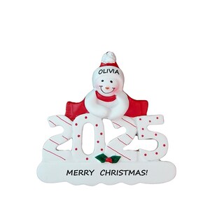 May include: A snowman Christmas ornament with the year 2025. The snowman wears a red hat and scarf, with the name "OLIVIA" on the hat. The numbers are white with red stripes and dots, and the base reads "MERRY CHRISTMAS!"