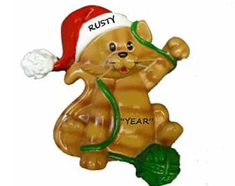 May include: A brown and orange cat wearing a red Santa hat with the name "RUSTY" on it. The cat is holding a green ball of yarn and the text "YEAR" is on the cat's body.