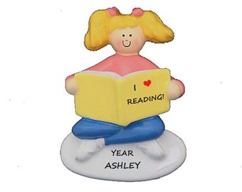 May include: A figurine of a girl with blonde pigtails wearing a pink shirt and blue pants, reading a yellow book. The book reads "I ❤️ READING!" and the base says "YEAR ASHLEY".