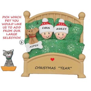 May include: A Christmas ornament featuring a bed with two figures wearing Santa hats, labeled "Chris" and "Ashley." A dog named "Aspen" is on the bed. The ornament has the text "Christmas 'Year'" and a heart.