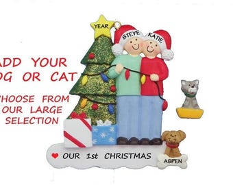 May include: A Christmas ornament featuring two figures in Santa hats, holding lights, with a Christmas tree, gifts, and pets. Text reads "ADD YOUR DOG OR CAT" and "OUR 1st CHRISTMAS". The ornament includes the names "STEVE" and "KATIE".
