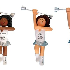 May include: Three personalized twirl girl ornaments. Each figurine has a different hair color and skin tone. The ornaments are wearing white tops with the text "TWIRL GIRL" and a name. They are wearing silver skirts and holding batons.