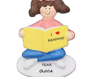 May include: A figurine of a girl with brown hair and pink bows, wearing a pink shirt and blue pants, reading a yellow book. The book reads "I ❤️ READING!" and the base says "YEAR OLIVIA".