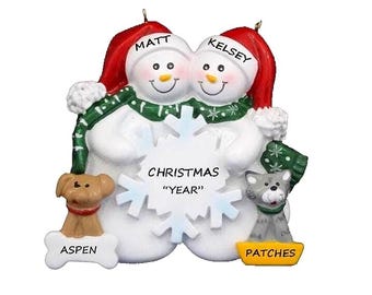 May include: A Christmas ornament featuring two snowmen wearing red Santa hats, green scarves, and holding a snowflake. The ornament includes a dog named Aspen and a cat named Patches. The text on the ornament reads "Christmas" and "Year".