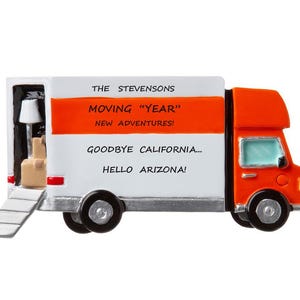 May include: A decorative moving truck ornament with an orange cab and white trailer. The trailer has an orange stripe and text that reads "The Stevensons, Moving 'Year', New Adventures!, Goodbye California... Hello Arizona!" The truck is loaded with boxes and a lamp.
