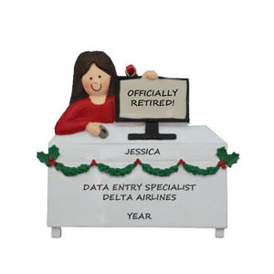 May include: A white desk ornament featuring a woman in a red top, holding a mouse. A computer monitor displays "OFFICIALLY RETIRED!". The desk is decorated with garland and text that reads "JESSICA", "DATA ENTRY SPECIALIST DELTA AIRLINES", and "YEAR".
