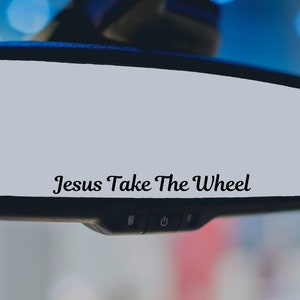 May include: A car's rearview mirror with the text "Jesus Take The Wheel" in a cursive font. The mirror is rectangular with rounded corners and a black frame. The background is blurred, suggesting the inside of a vehicle.