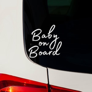 K&ouml;nnte beinhalten: Wei&szlig;er Autoaufkleber mit den Worten "Baby on Board" in einer Schreibschrift. Der Aufkleber befindet sich auf der Heckscheibe eines wei&szlig;en Autos, darunter ist das rote R&uuml;cklicht zu sehen.