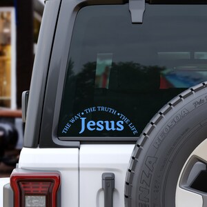 Puede incluir: Un coche blanco con una pegatina azul y blanca en la ventana trasera que dice "Jes&uacute;s, El Camino, La Verdad, La Vida".