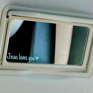 May include: A white car visor with a mirror that has the words "Jesus loves you" written in blue with a small blue heart.